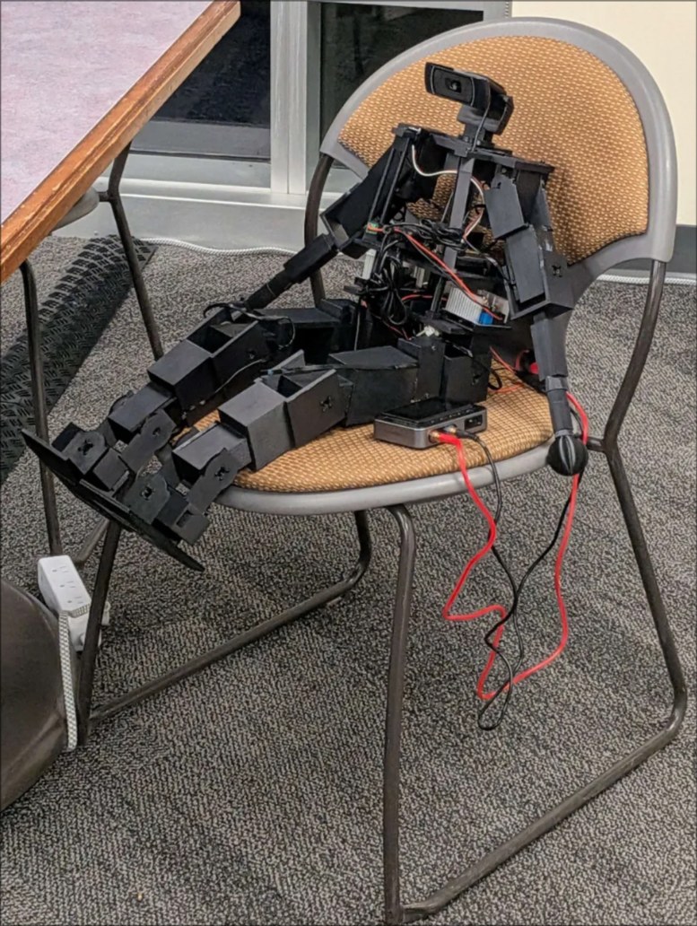 Proto, a black 3D printed humanoid robot, seated on a chair with a camera mounted where its head would be.